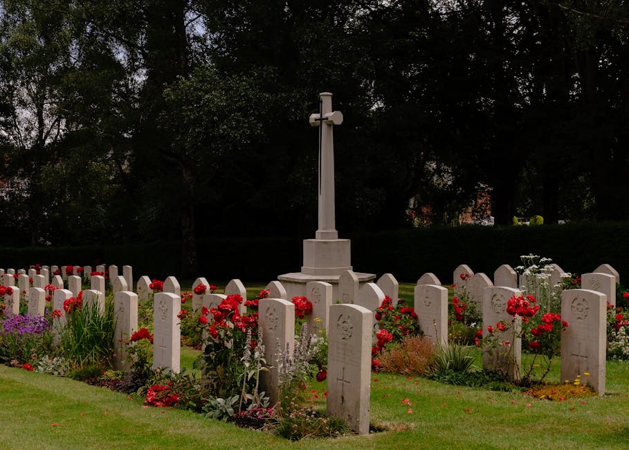 A neatly maintained cemetery border features a circular arrangement of white gravestones with decorative carvings, set amidst lush green grass. Bright red and purple flowers are planted along the edges of the graves, adding color and vibrancy to the scene. In the center of the arrangement, a tall white cross monument stands on a tiered base, surrounded by rich, moist soil. Behind the border, a line of dark green trees provides a shaded backdrop, with dappled sunlight filtering through the foliage. The overall scene showcases careful garden maintenance and landscaping work by local gardeners, with trimmed grass, healthy flowering plants, and a clean, orderly layout, demonstrating ongoing lawn care and hedge trimming efforts by Gardeners Brompton.