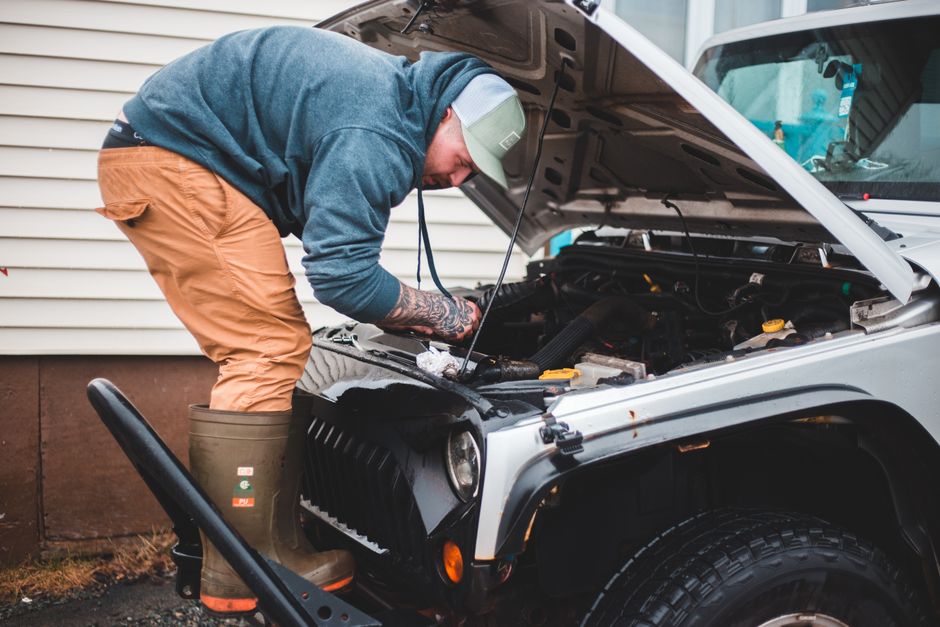 A man wearing a grey cap, a blue hoodie, and khaki pants is working on a silver off-road vehicle parked outdoors. He is bent over, inspecting or repairing the engine with the hood open, revealing various engine components and hoses. The man has tattooed arms visible as he uses a tool, and he is standing on a patch of gravel or dirt beside a garden wall with horizontal white siding. Nearby, a black garden tool with a curved handle rests on the ground. The setting suggests a practical outdoor workspace, possibly related to garden maintenance or landscaping work, with natural light illuminating the scene. The focus is on the man performing vehicle repairs, which might relate to transport or equipment maintenance for outdoor garden services, aligning with themes discussed on the Brompton garden services access problems and fixes page from Gardeners Brompton.